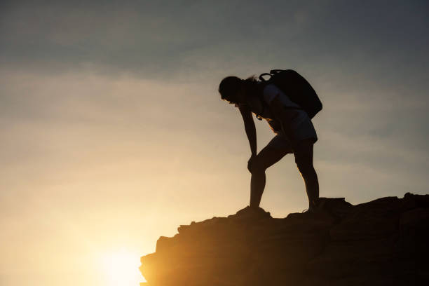 Silhouette Tired hiking  woman resting and standing on top of cliff hill during sunset after climbing to mountain summit peak with copy space for text. Success to goal. Exhausted sport work out and Healthy lifestyle.