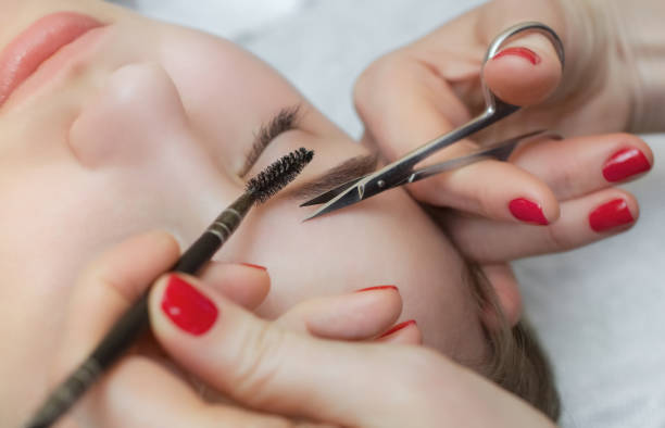 Master makeup corrects, gives shape to and cut with scissors eyebrows in a beauty salon. Professional care for face.