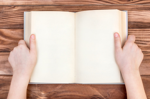 Child's hands holding opened blank book over wooden table. Top view.