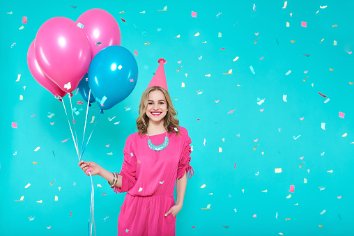 Gorgeous birthday girl in party outfit holding colourful balloons. Attractive trendy teenager celebrating birthday. Party and flying confetti on pastel blue background.