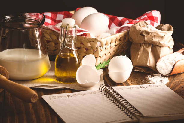 Ingredients of a recipe, with eggs, milk, oil and wheat flour, upon a rustic wooden table, and a notebook.