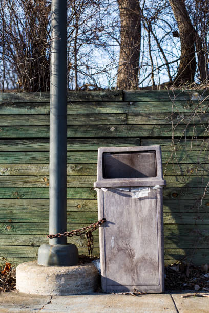 istock Grey garbage can chained to post in parking lot with wood wall, tree branches and other debris in spring 941953902
