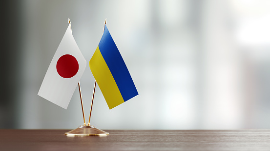 Japanese and Ukrainian flag pair on desk over defocused background. Horizontal composition with copy space and selective focus.