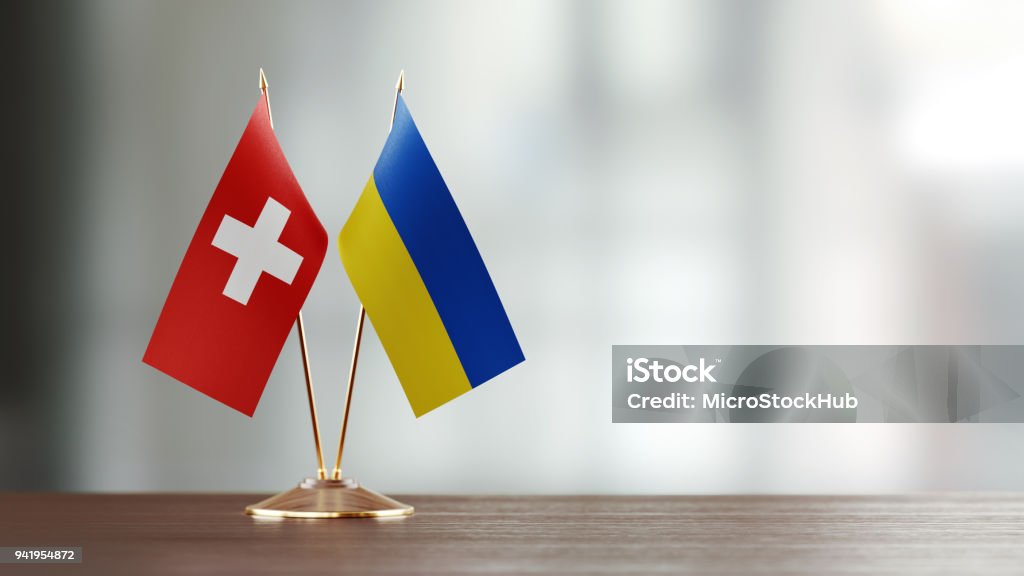 Swiss And Ukrainian Flag Pair On A Desk Over Defocused Background Swiss and Ukrainian flag pair on desk over defocused background. Horizontal composition with copy space and selective focus. Ukraine Stock Photo