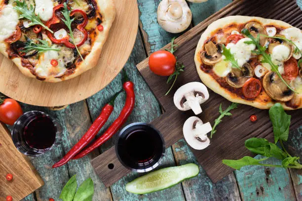 Pizza party flatlay. Rustic pizzas with bell pepper, mushrooms and arugula served with raw chili pepper, champignons, tomatoes and red wine on shabby blue background overhead view