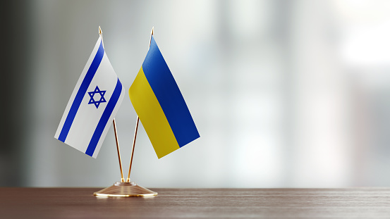 Israeli and Ukrainian flag pair on desk over defocused background. Horizontal composition with copy space and selective focus.