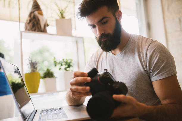 istock Good looking bearded man looking photos on a camera while sitting in a coffee shop doing work on a laptop. 941992224