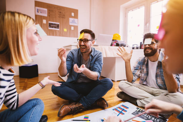 Pretty young designer coworkers enjoying free time and raising togetherness and team spirit while sitting in a circle on the floor of the office and playing games with paper stickers on the forehead. Pretty young designer coworkers enjoying free time and raising togetherness and team spirit while sitting in a circle on the floor of the office and playing games with paper stickers on the forehead. office fun business adhesive note stock pictures, royalty-free photos & images