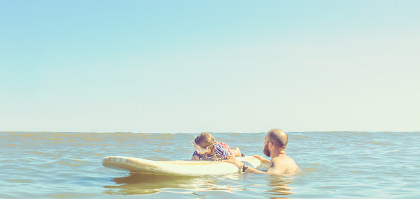 Dad holds a surfboard with a child on it in a candid moment, teaching surfing