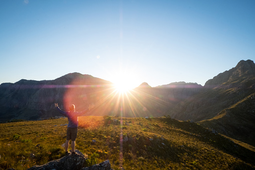 A young male hiker stands with his arms raised on a rock looking at the sunrise over the mountains, Cape Town South Africa