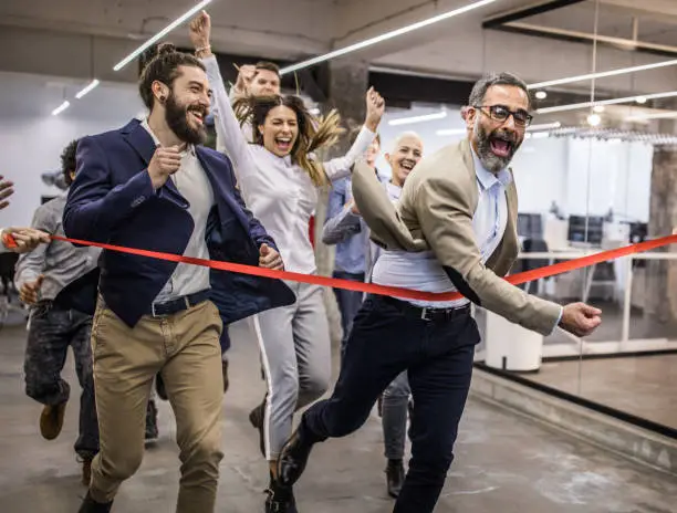 Large group of business people having fun while crossing the finish line at casual office. Focus is on young bearded man.