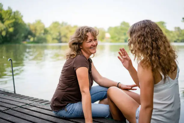 Photo of Cheerful mother and daughter sitting on the edge of a wooden jetty