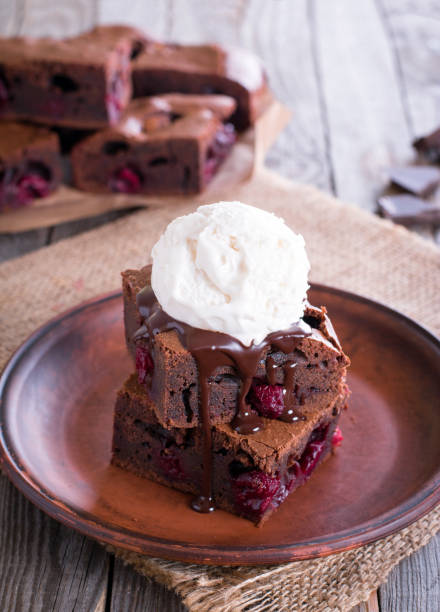 Chocolate brownie square pieces in stack on white plate decorated with mint leaves and cocoa powder on white vintage wooden background. American traditional delicious dessert. Close up photography.