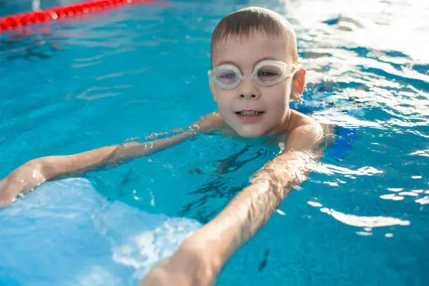 Cheerful cute kid in swimming googles learning to swim and using kickboard for floating in pool
