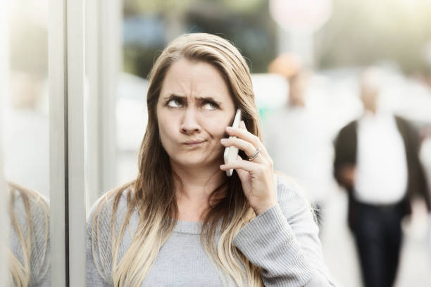 Frustrated woman in street listening on phone rolls her eyes A blonde woman standing in the street listens to something - possibly a recorded message - on her mobile phone and rolls her eyes, grimacing in frustration. rolling eyes irritation displeased facial expression stock pictures, royalty-free photos & images