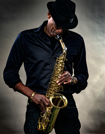 Musician with saxophone against a gray backdrop. Men plays the saxophone, wearing a black hat and clothes