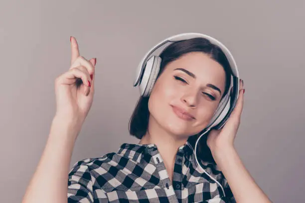 Photo of A horizontal image of young cute happy woman in checkered shirt listening to her favourite track and pointing up