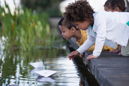 Three kids playing competition with paper boats at a pond.