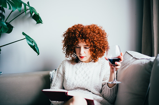 Redhead young woman with curly hair, sitting on sofa, reading a book and drinks red wine (toned image, selective focus)