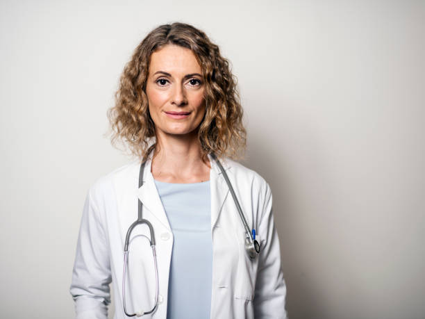 Portrait of female doctor against wall in hospital Portrait of confident smiling female doctor. Mid adult medical professional is wearing lab coat and stethoscope. She is standing against gray wall in hospital. female doctor photos stock pictures, royalty-free photos & images