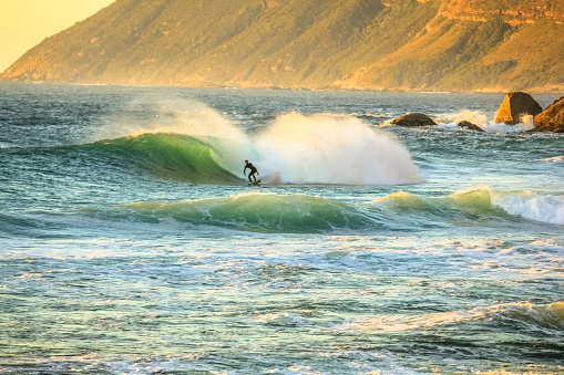 Noordhoek Beach at sunset. Surfing in Cape Town, South Africa. Surfer tries to take a high and powerful wave. Atlantic coast in Table Mountain National Park. Extreme sports leisure concept.