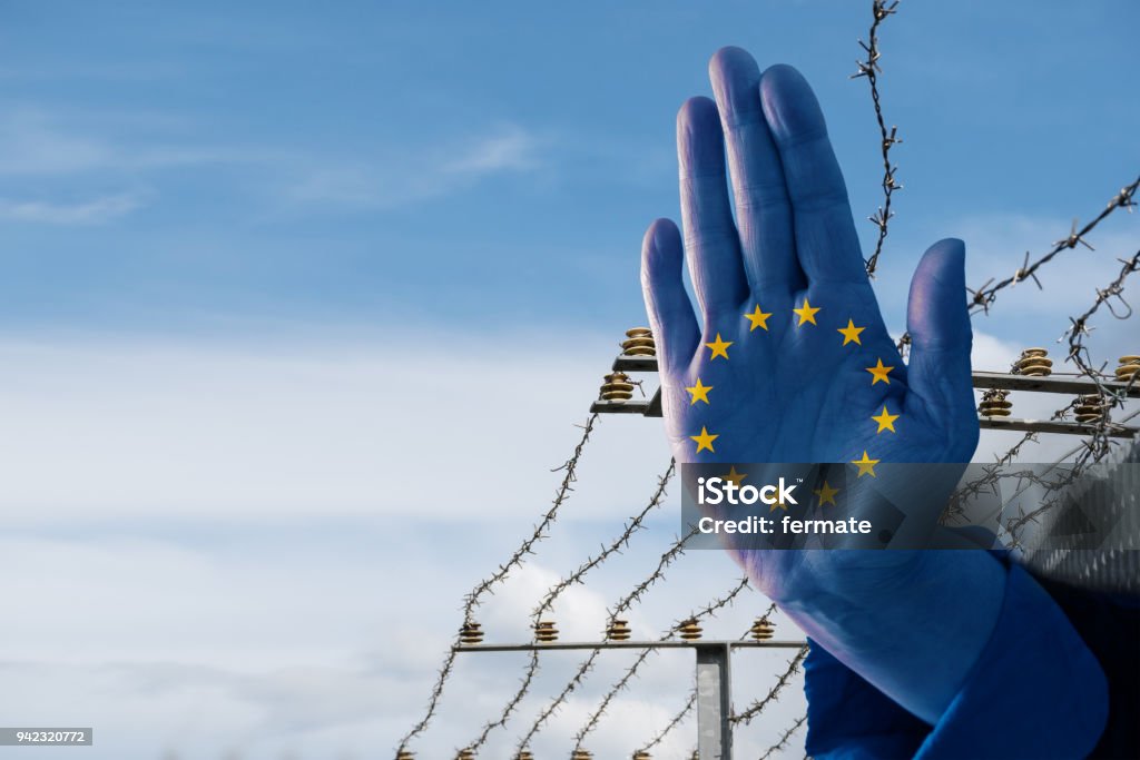 Hand with European flag stops immigration of refugees, blurred border fence in the background, blue sky with copy space Schengen Agreement Stock Photo