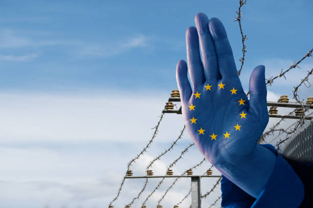 Hand with European flag stops immigration of refugees, blurred border fence in the background, blue sky with copy space stock photo