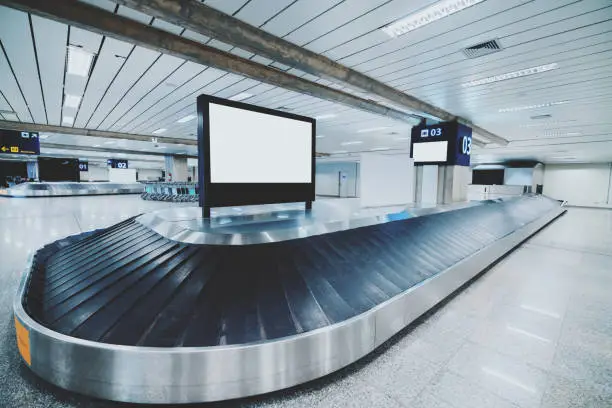 Wide-angle view of a modern baggage conveyor belt in a contemporary airport terminal: multiple ribbon moving sections, chromed body, empty white informational LCD screen template on the top