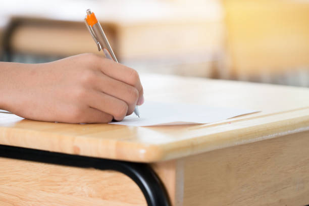 istock Education test concept : Hands with pen over application form, Students taking exams, writing examination room with undergraduate inside. Student sitting learning lessons doing final in classroom 942443496