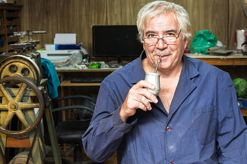 Portrait of a Argentinian jeweler wearing a blue smock and drinking mate in his jewerly workshop.