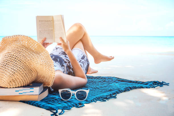 frau liest ein buch am strand in freizeit sommerurlaub - sonnenbaden stock-fotos und bilder