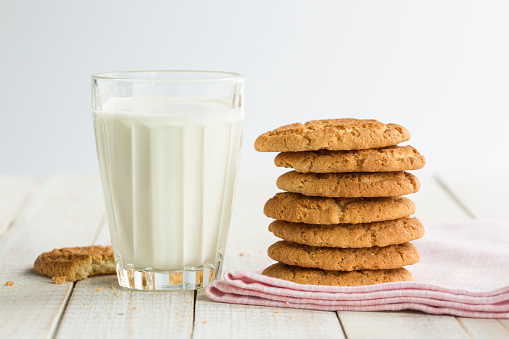 Stack of homemade cookies (vanilla, butter, oat or similar) on a pale pink cloth napkin on white rustic wooden surface (table) with a glass of milk. One of the cookies is bitten. Idea of snack for kids. No people. Horizontal orientation photography. Front view. White background. Studio photo-shot using natural day light. This image evokes the warmth of childhood home, the cookies of your mother or your grandmother.