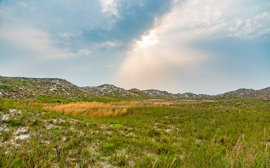 Sunlight breaking through clouds behind grass covered sand dunes.