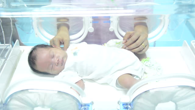 Close up of mother taking care of newborn baby inside incubator, mother looking at newborn baby sleeping inside medical intensive care units incubator
