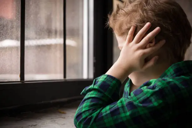 Photo of Little boy standing behind the window in sad mood. Sad Teenager looking in the Window and closing his ears with hands. Unhappy child in a plaid shirt. Alone at home. Upset.