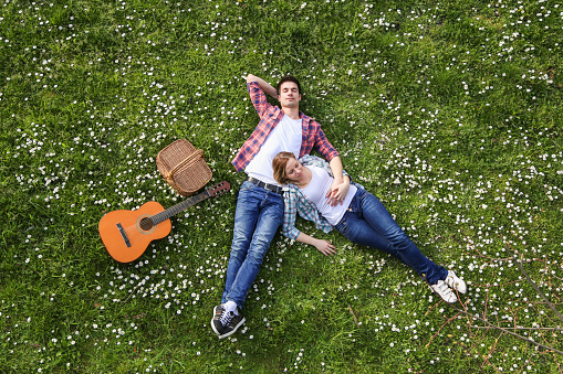 Happy attractive heterosexual couple enjoying picnic time on lawn with acoustic guitar. Both Caucasian in casual clothing, about 20-25 years old