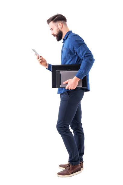 Professional business male executive checking phone carrying tablet and laptop under arm. Side view. Full body isolated on white background.