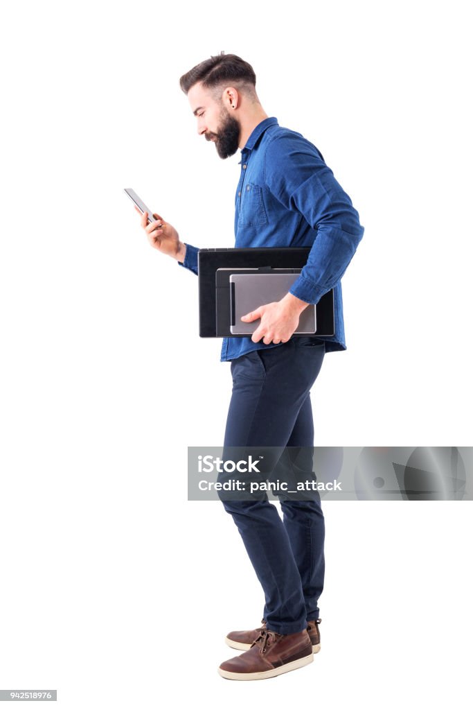 Profissional masculino executivo verificando o telefone carregando tablet e computador portátil debaixo do braço. Vista lateral. - Foto de stock de Fundo Branco royalty-free