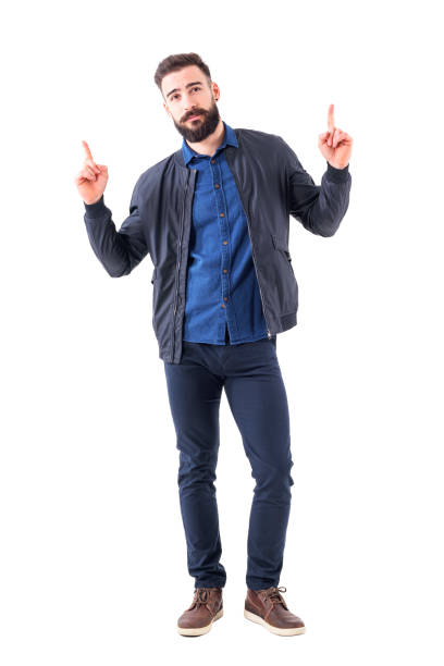Cool relaxed bearded man in blue bomber jacket pointing fingers up above and looking at camera