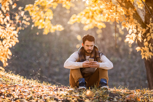 Young man relaxing in autumn day in nature and typing text message on mobile phone.