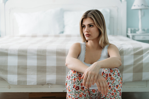 Depressed young woman sitting on bed at home alone