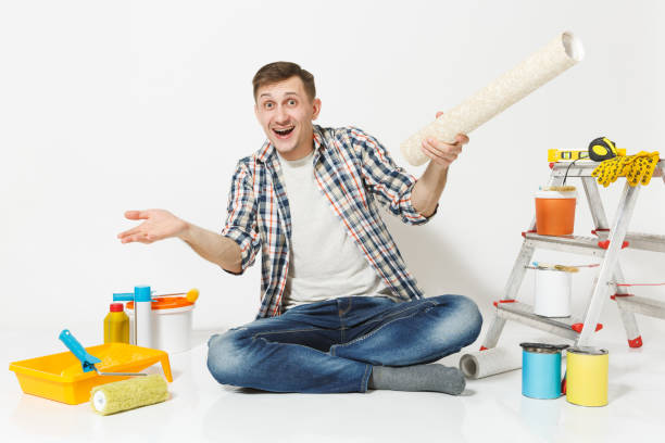 istock Smiling man spreading hands, sitting on floor with rolls of wallpaper, instruments for renovation apartment room isolated on white background. Gluing accessories, painting tools. Repair home concept. 942534698