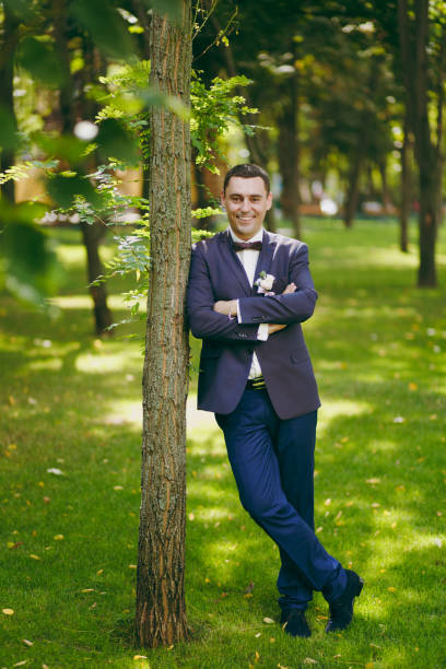 Beautiful wedding photosession. Handsome smiling groom in blue formal suit, white shirt, bow tie and boutonniere of flowers and ribbons near tree on a wedding walk in the big green park on sunny day Beautiful wedding photosession. Handsome smiling groom in blue formal suit, white shirt, bow tie and boutonniere of flowers and ribbons near tree on a wedding walk in the big green park on sunny day. niñas stock pictures, royalty-free photos & images