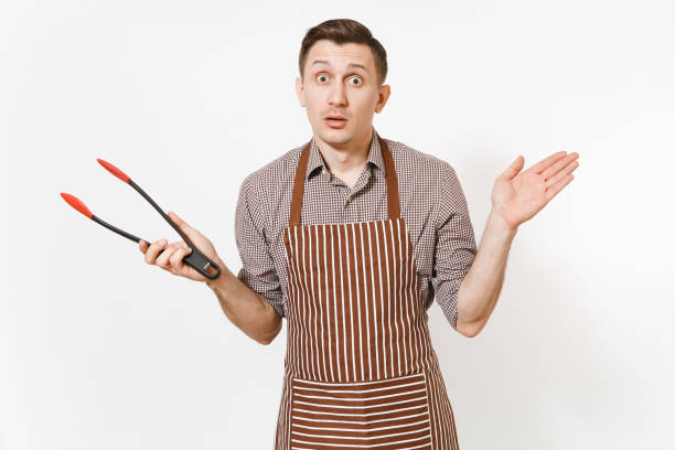 istock Young man chef in striped brown apron, shirt holding black kitchen serving plastic pair of tongs for salad isolated on white background. Male housekeeper or houseworker. Kitchenware, cuisine concept. 942537858