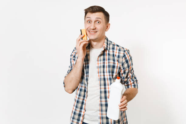 istock Young housekeeper man holding sponge, white blank empty cleaning bottle with cleaner liquid isolated on white background. Male doing house chores. Copy space for advertisement. Cleanliness concept. 942537882