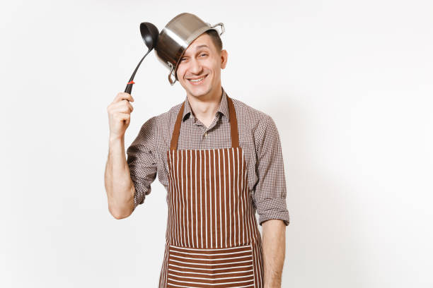 istock Fun mad man in striped apron with ladle, silver stainless glossy aluminium empty stewpan, pan or pot on head isolated on white background. Male housekeeper or houseworker. Kitchenware, dishes concept. 942538190