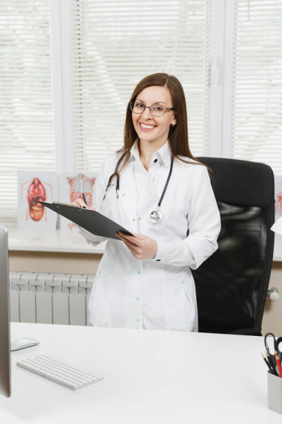 Young female doctor standing at desk, working with clipboard and medical documents in light office in hospital. Woman in medical gown, stethoscope in consulting room. Healthcare, medicine concept. Young female doctor standing at desk, working with clipboard and medical documents in light office in hospital. Woman in medical gown, stethoscope in consulting room. Healthcare, medicine concept niñas stock pictures, royalty-free photos & images