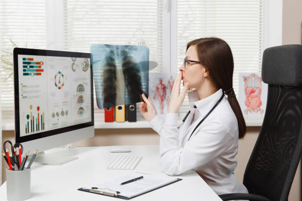 Female doctor sitting at desk, working on computer, holding X-ray of lungs, fluorography, roentgen in light office in hospital. Woman in medical gown, stethoscope in consulting room. Medicine concept. Female doctor sitting at desk, working on computer, holding X-ray of lungs, fluorography, roentgen in light office in hospital. Woman in medical gown, stethoscope in consulting room. Medicine concept niñas stock pictures, royalty-free photos & images