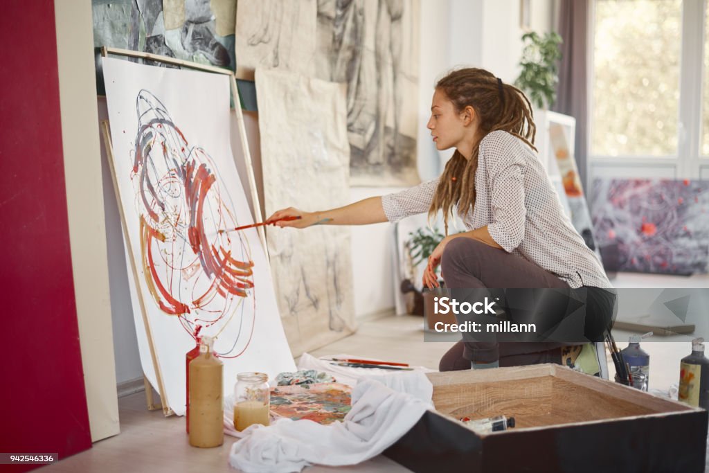 Artistic girl sitting in studio and paint on easel. Artistic girl sitting in studio and paint on easel while sitting on chair. In hand paint brush. Artist Stock Photo