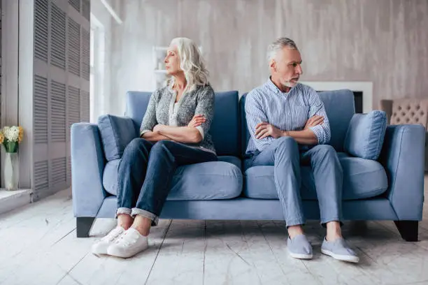 Senior couple at home. Handsome old man and attractive old woman are having relationship problems. Sitting on sofa together and looking to opposite sides.